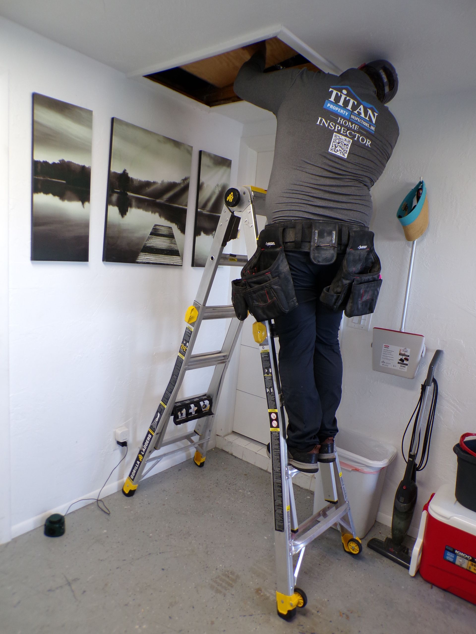Person on a ladder, working on an attic access in a white-walled room, wearing a tool belt and a gray Titan sweatshirt.