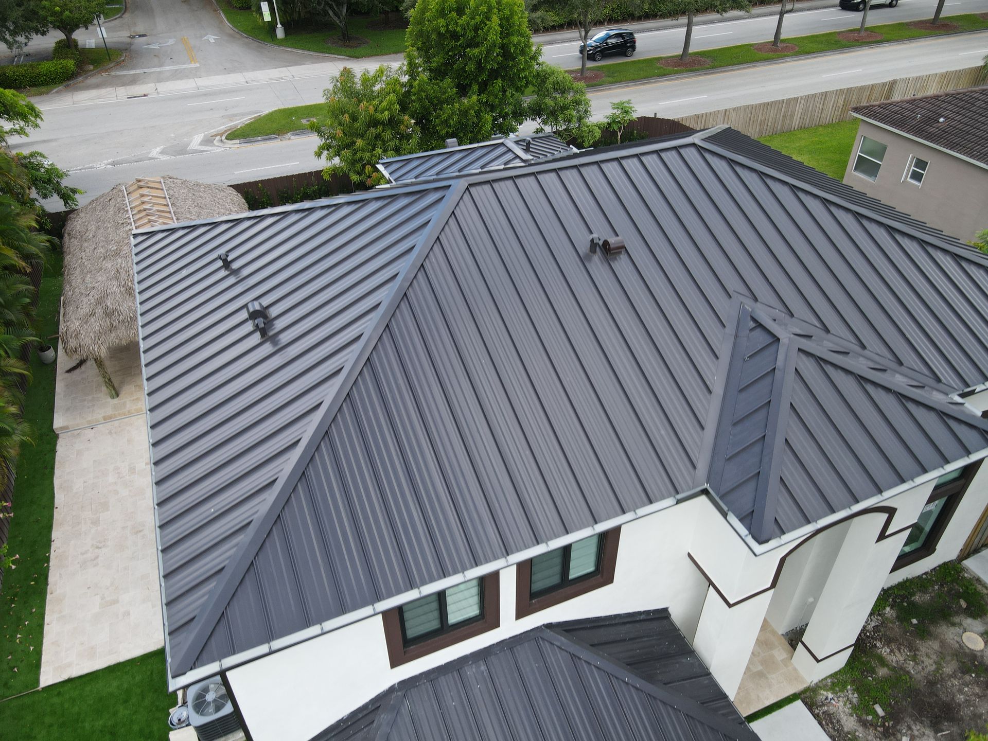 Dark gray metal roof on a white house with a circular driveway and small tiki hut.