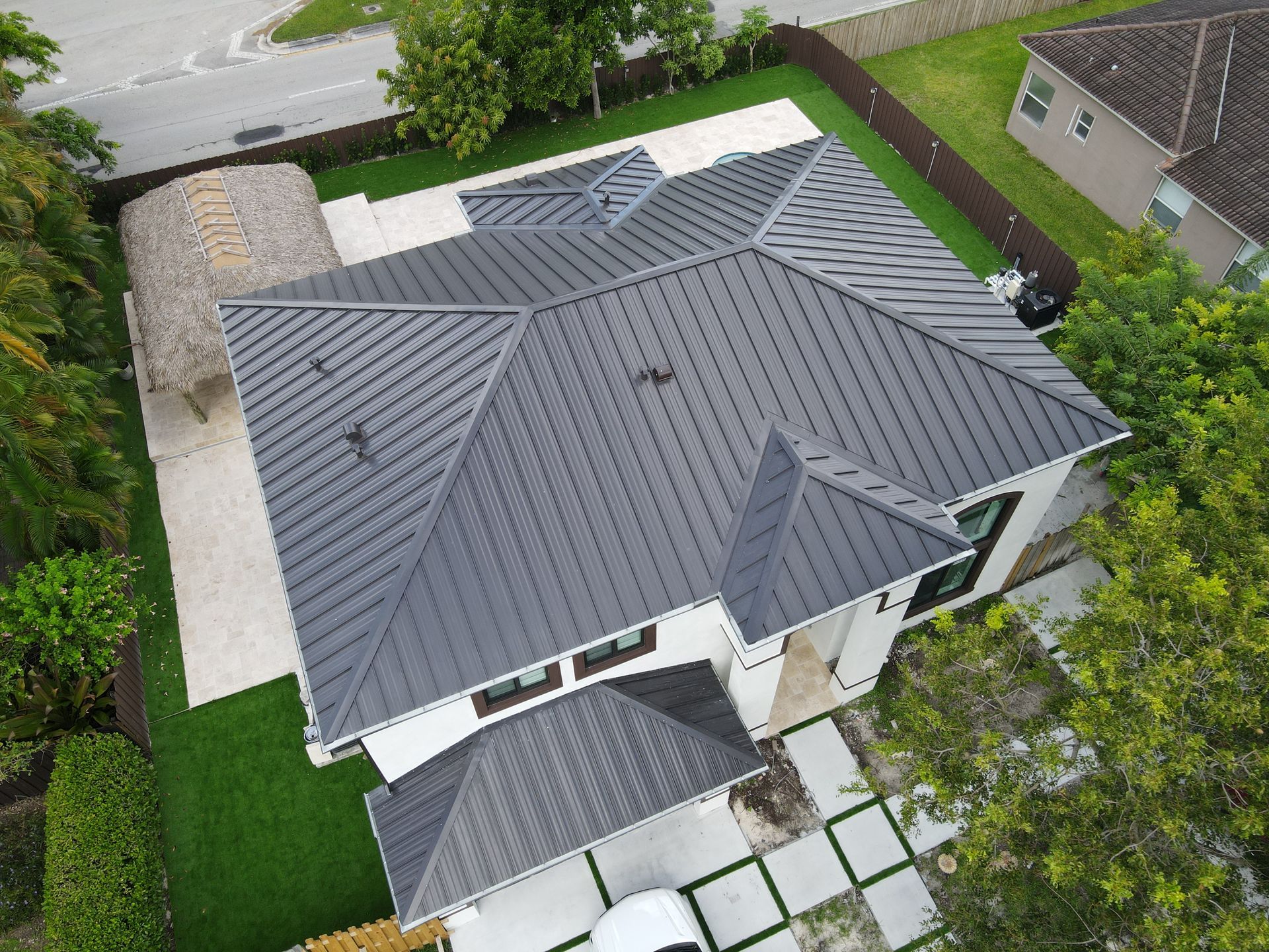 Aerial view of a modern house with a dark gray metal roof, surrounded by green lawn and trees.