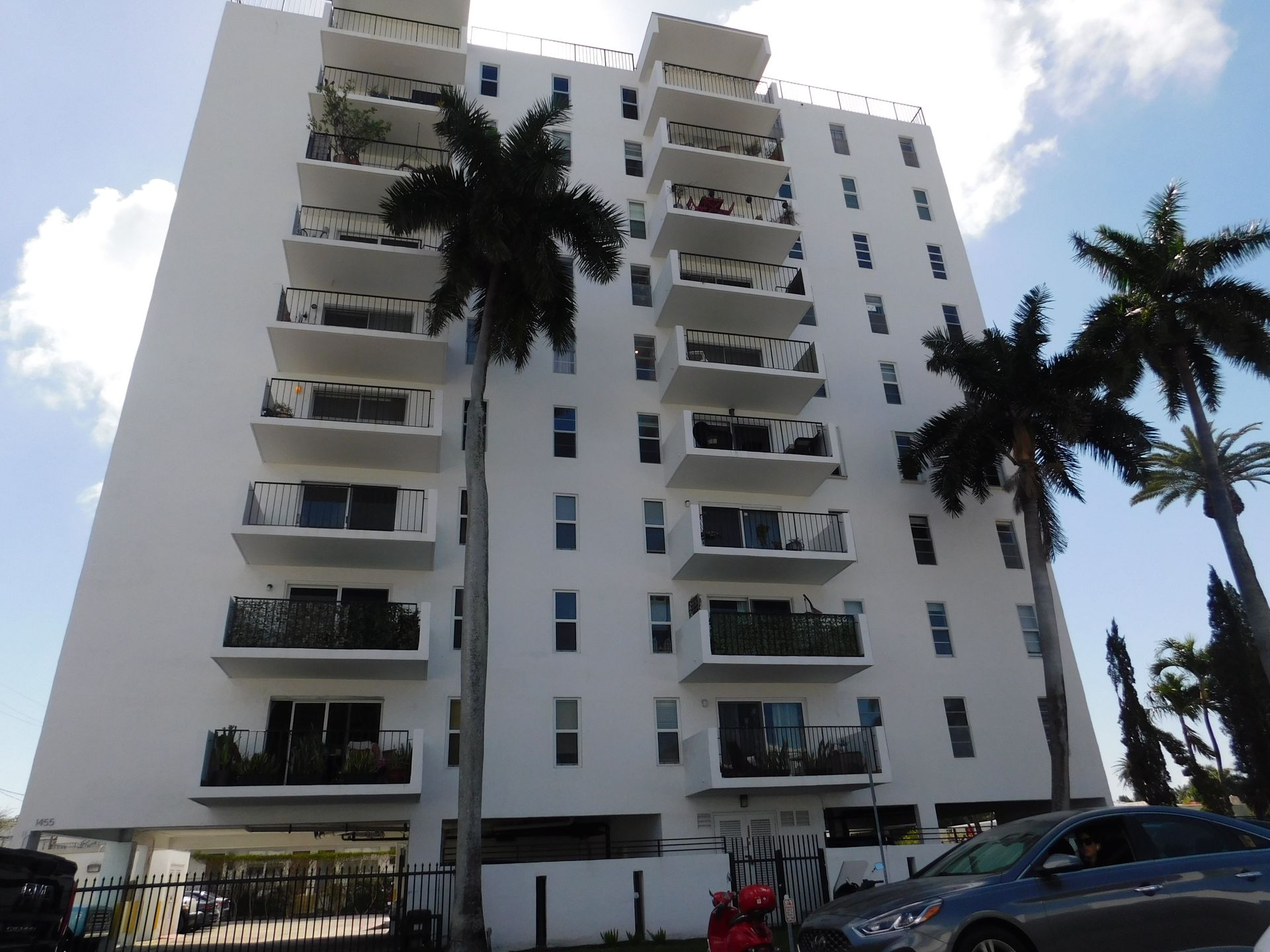 White apartment building with balconies and palm trees, a car parked nearby.