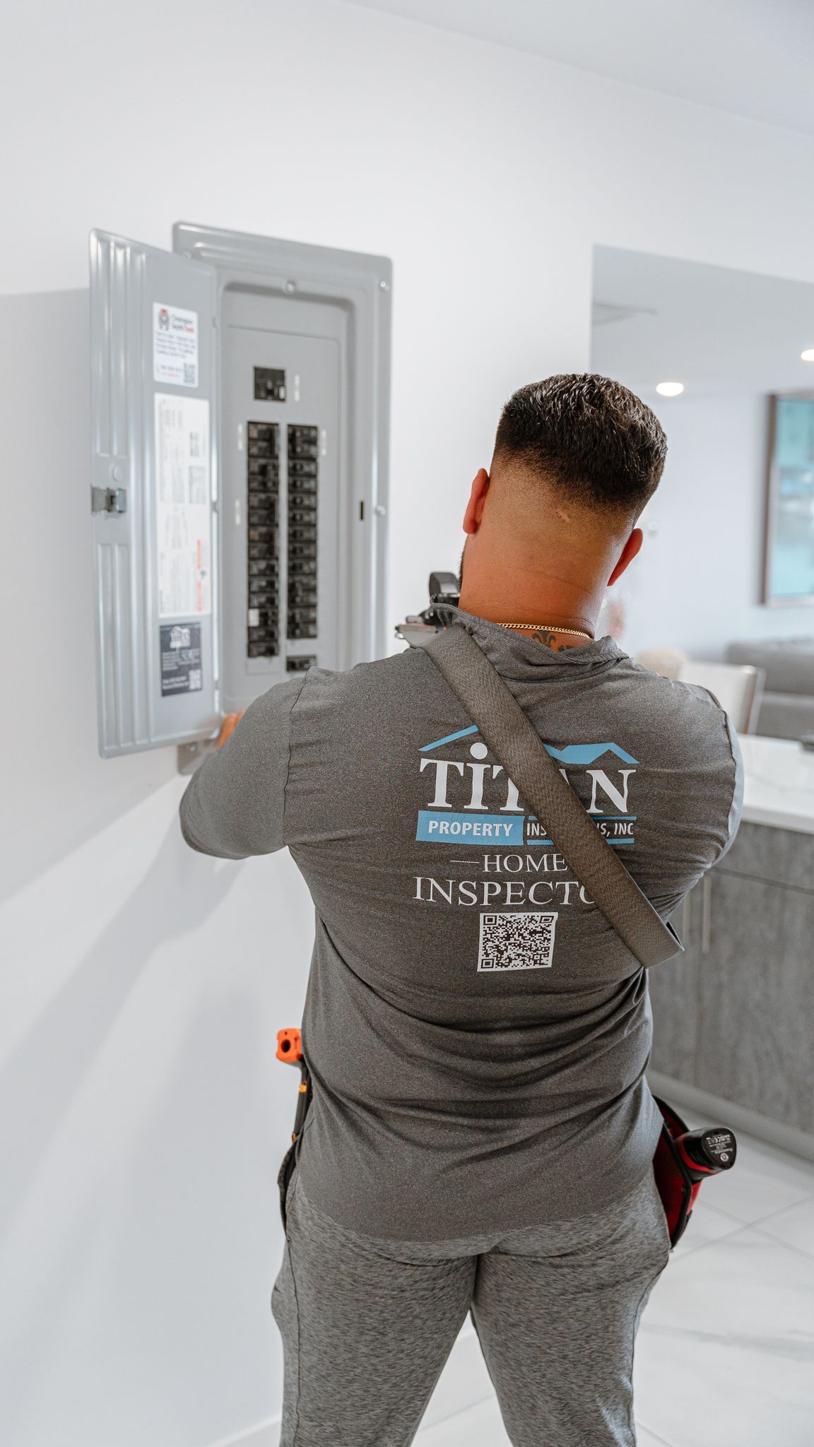 Electrician working on a circuit breaker panel on a white wall. He is wearing a grey shirt with a logo, in a modern home.