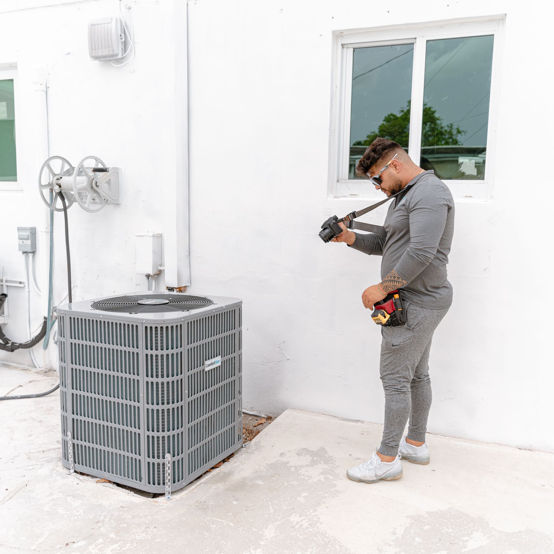 Man in gray sweatsuit photographing an AC unit outside a white building.