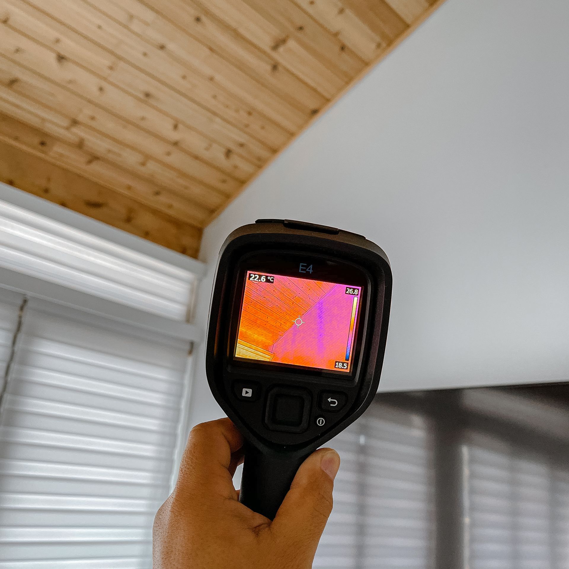 Hand holding thermal camera pointed at white ceiling; wood and blinds visible.