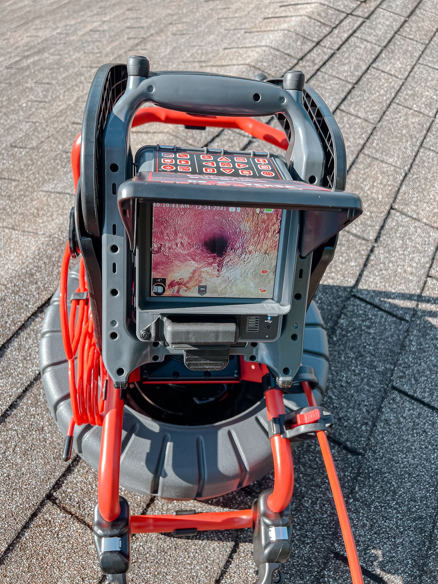 A plumbing inspection camera on a rooftop, showing a monitor displaying pipe interior. Red and black colors.