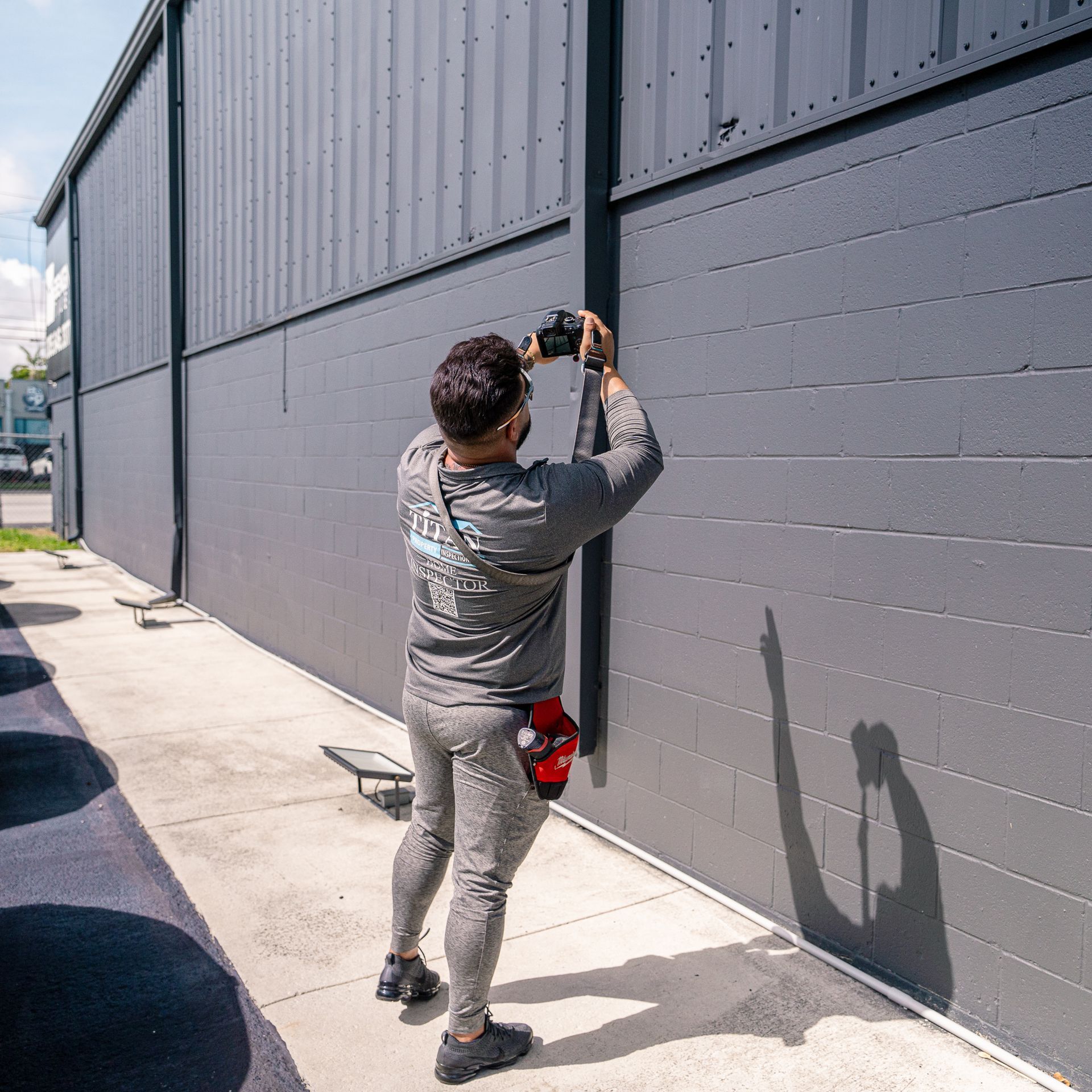 Man taking photo of a gray building. He is wearing gray clothing and sunglasses. The setting is outdoors.