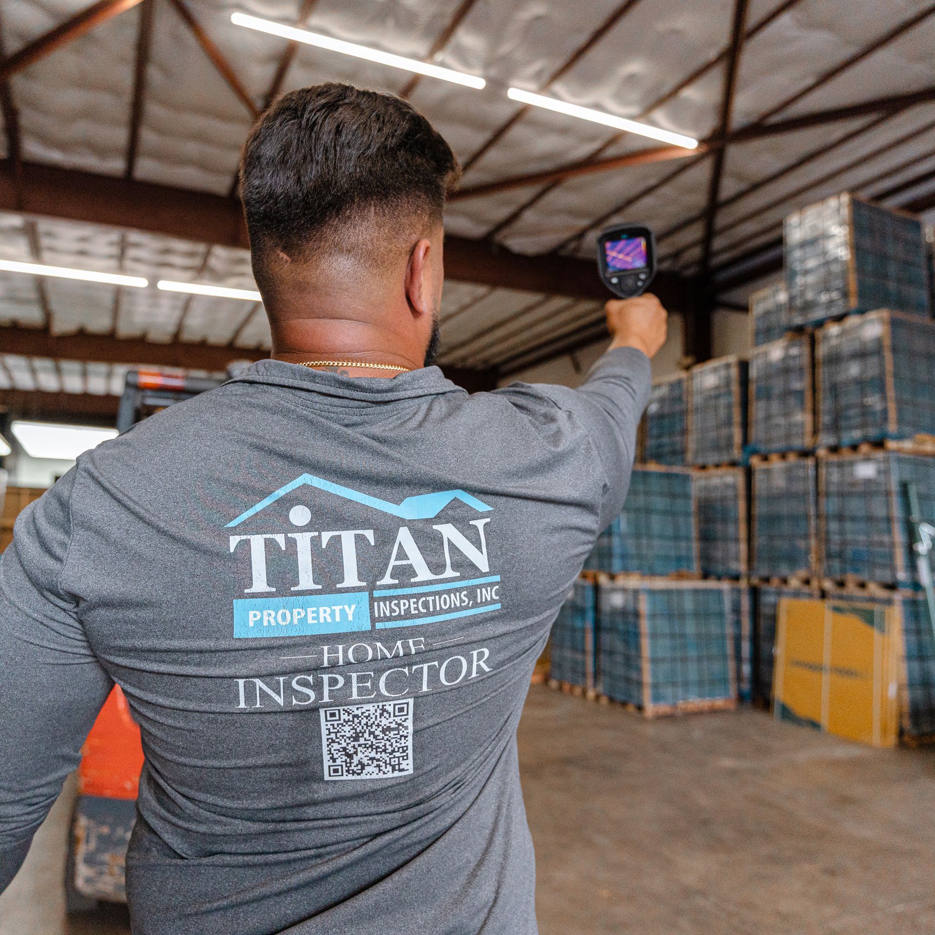 Man in gray shirt holding a thermal camera inspecting stacks of materials in a warehouse.