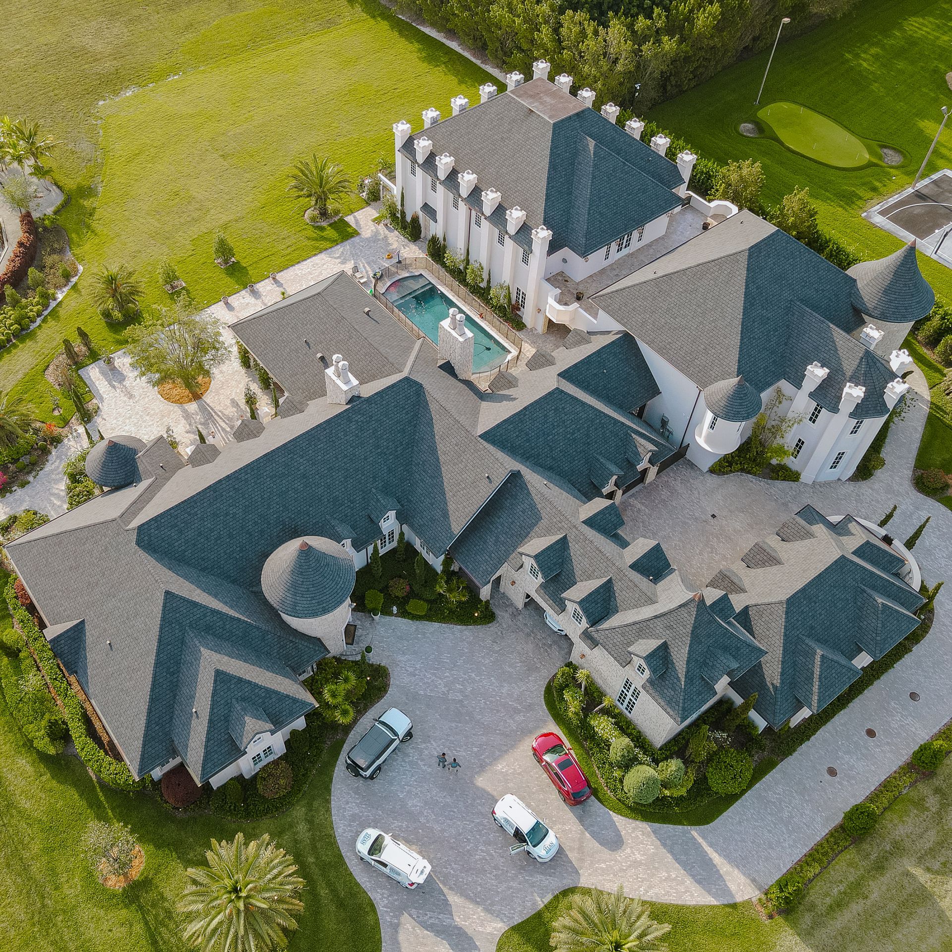 Aerial view of a large, luxurious mansion with multiple roof sections, a swimming pool, and parked cars on a paved driveway.