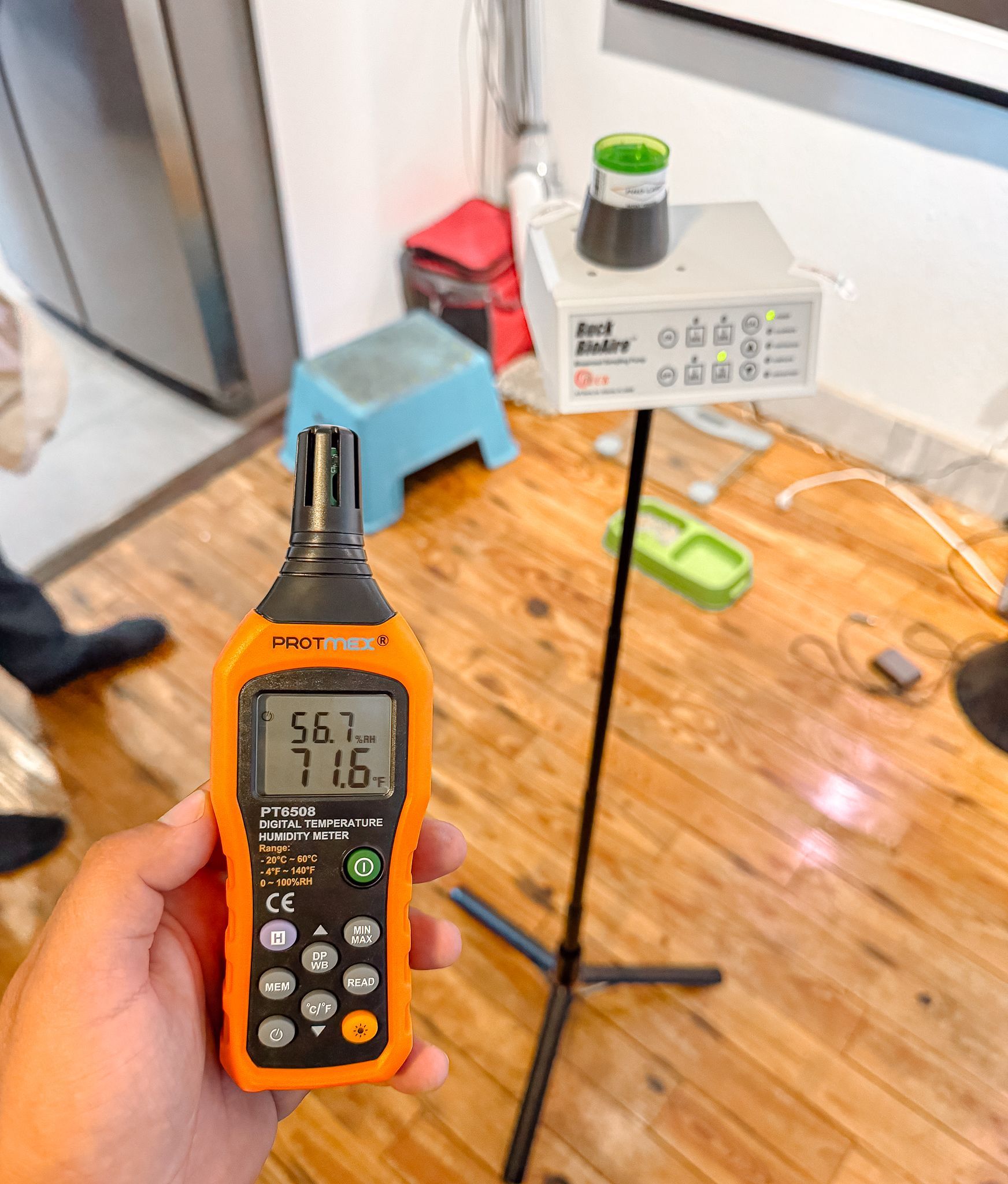 Hand holding orange meter displaying humidity, temperature near a white air quality monitor on a stand. Indoors.