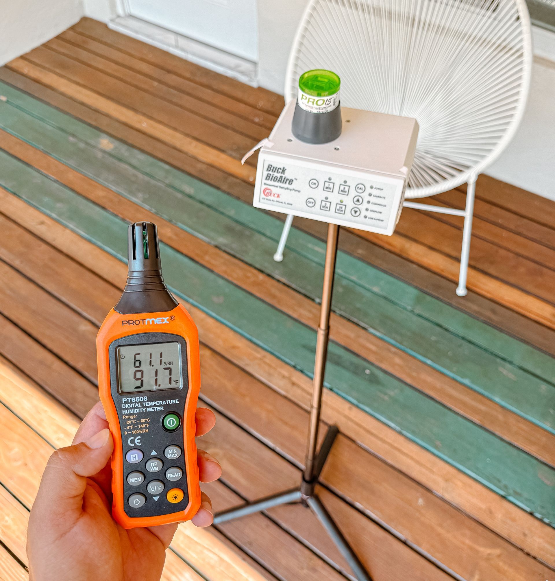 Person holds orange meter displaying 91.7. Beside them is white stand with device and a green-topped container on a wooden deck.