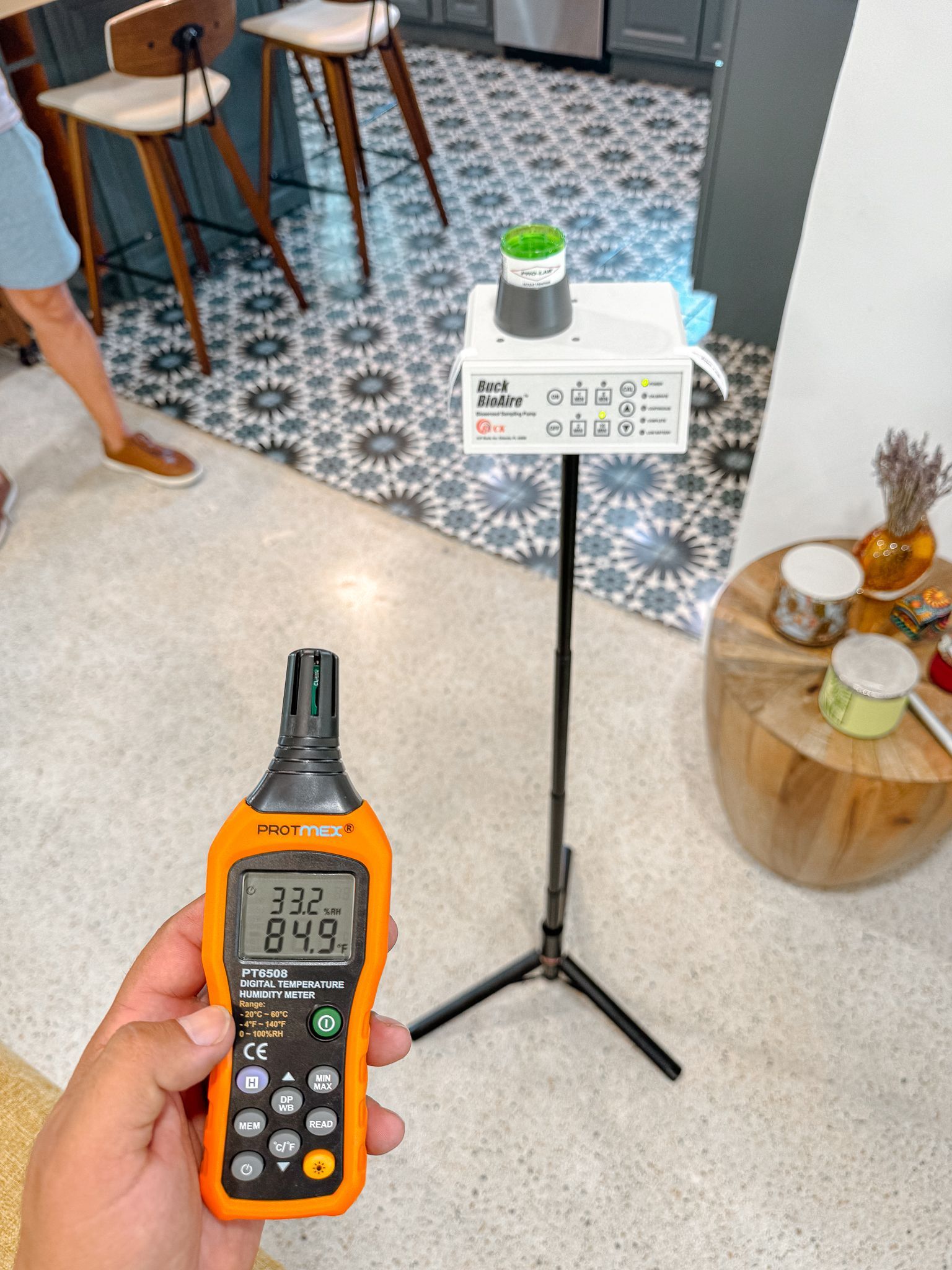A person holds an orange meter. A white device sits on a tripod. Blue and white tiled floor.