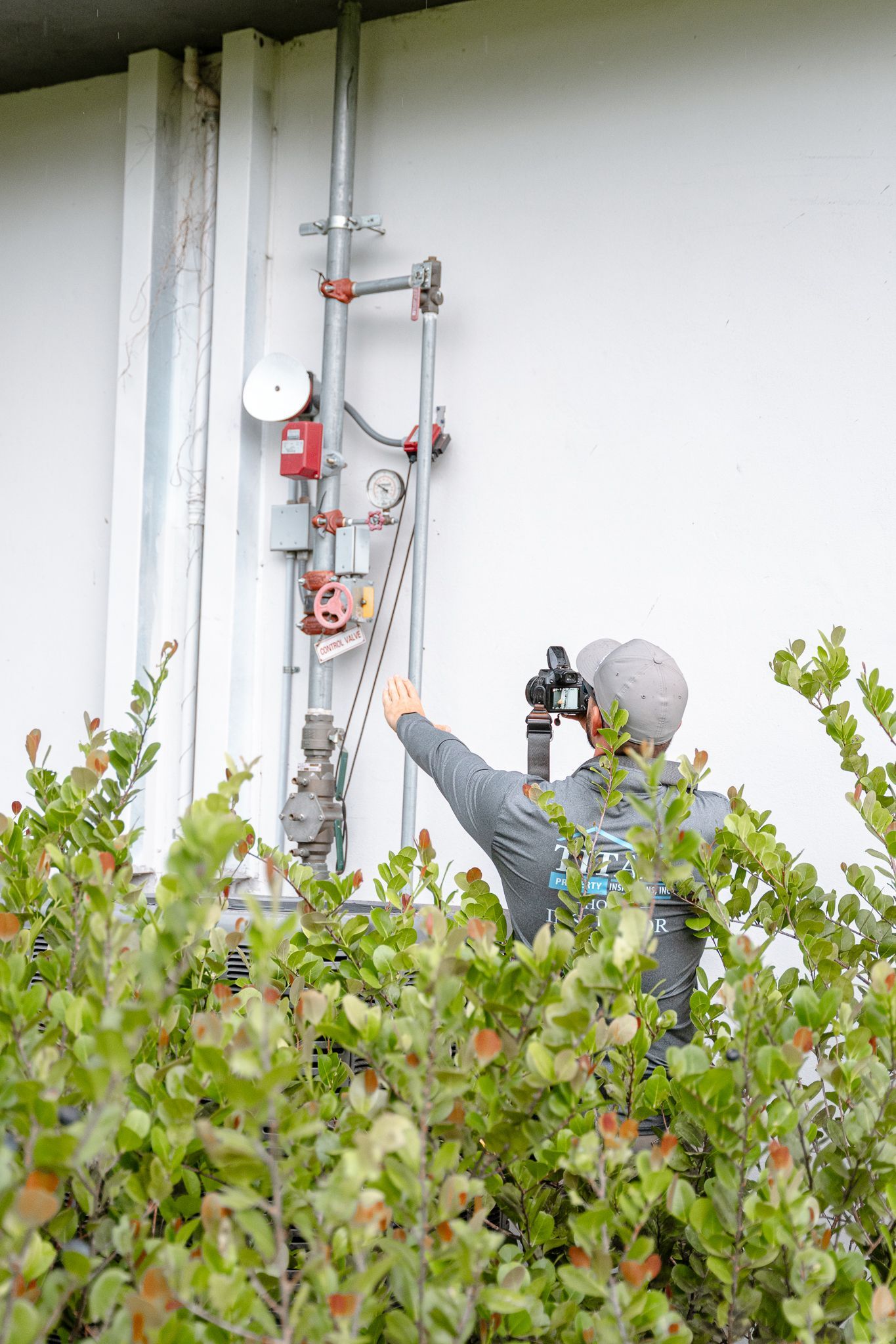 Person points at pipes and equipment on a white building wall, framed by green bushes.
