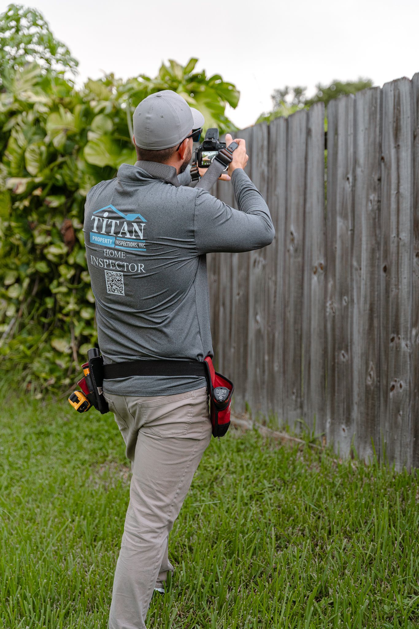 Person in grey shirt and cap taking photo of a fence with a camera in a yard.