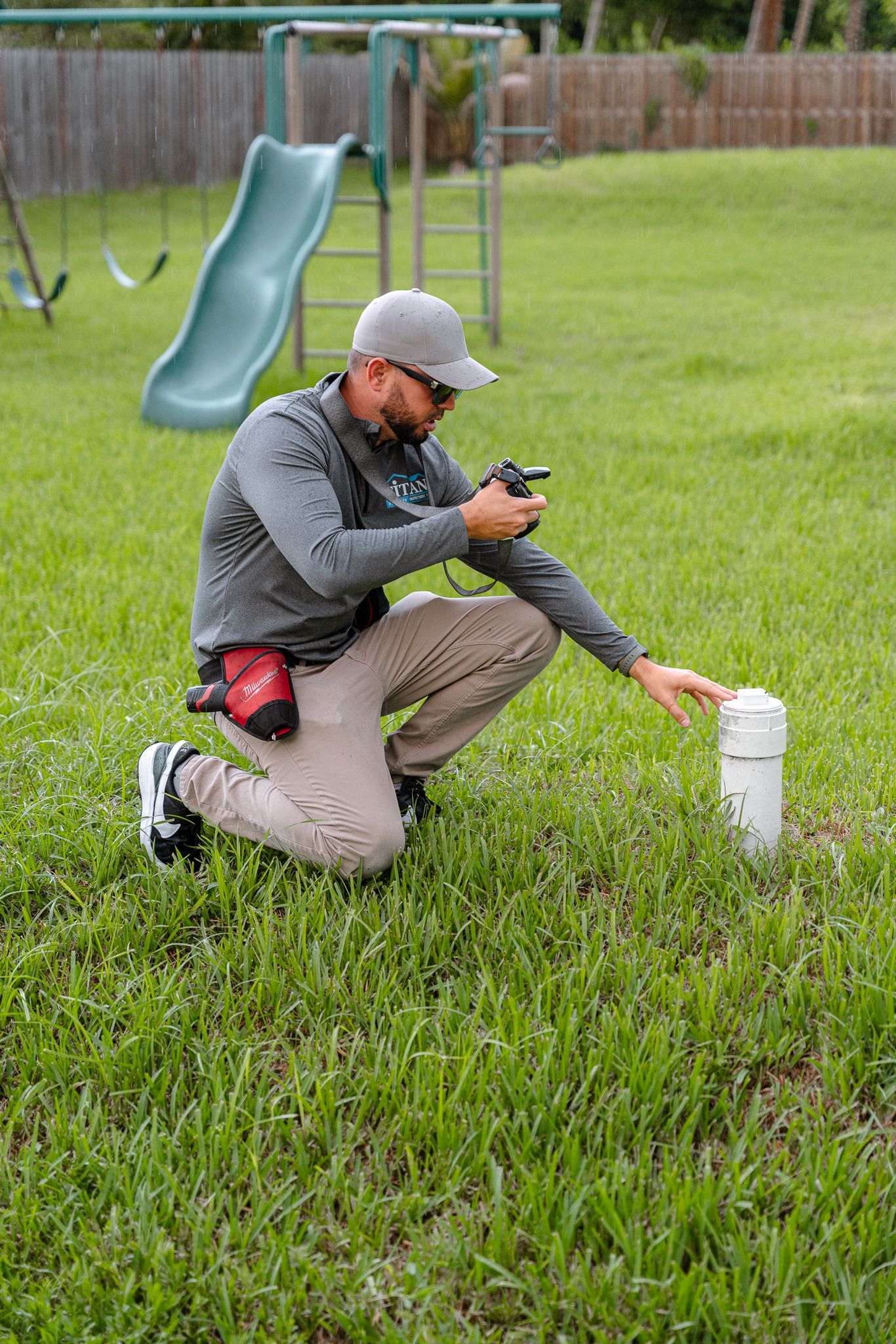 Man kneeling in a yard, examining a pipe with a device. A playground is in the background.