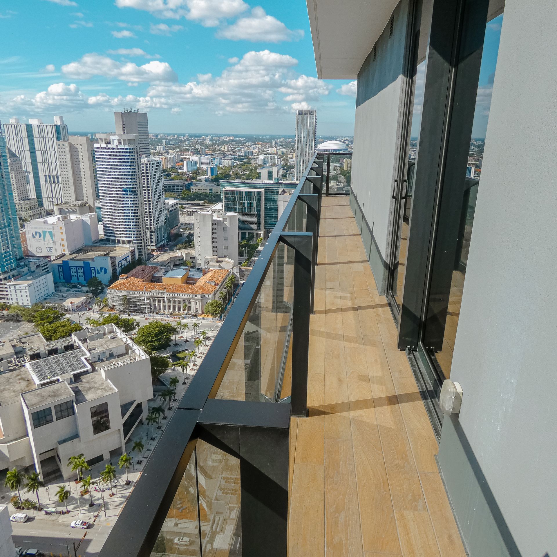 Balcony with city view. Wooden floor, glass railing, tall buildings, and blue sky with clouds.