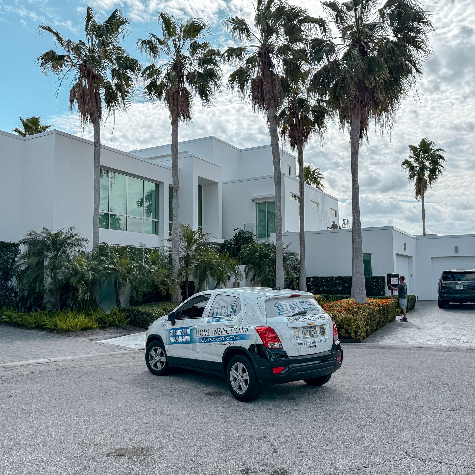 White SUV with company logo parked in front of a modern white house with palm trees.