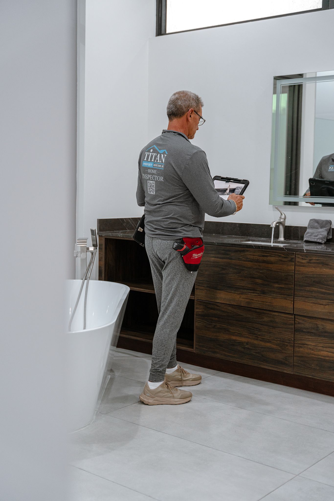 Man inspecting a modern bathroom, holding a tablet. Features a large vanity, tub, and window.