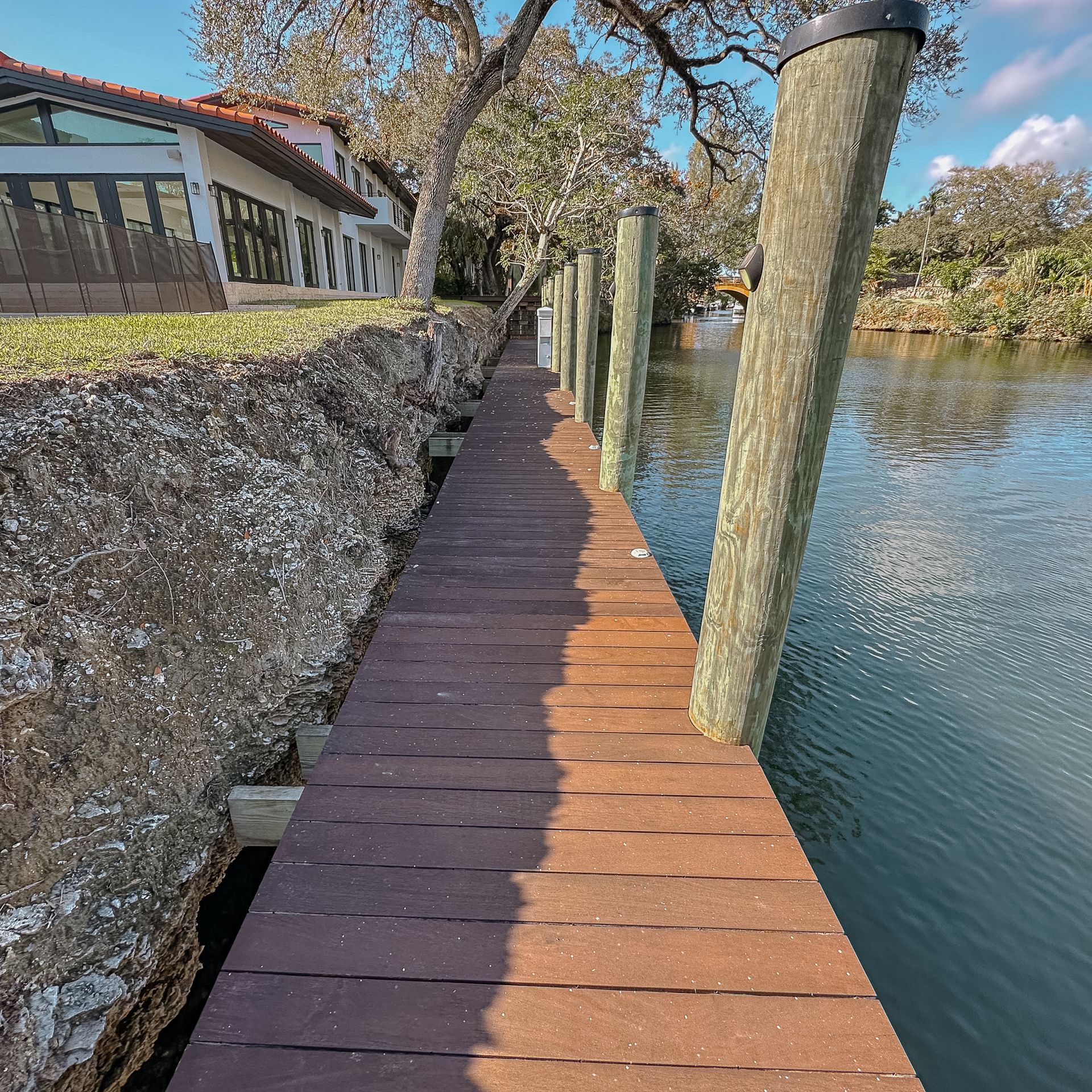 Wooden walkway next to water, with house in the background. Brown planks, weathered posts, and rocky shoreline.