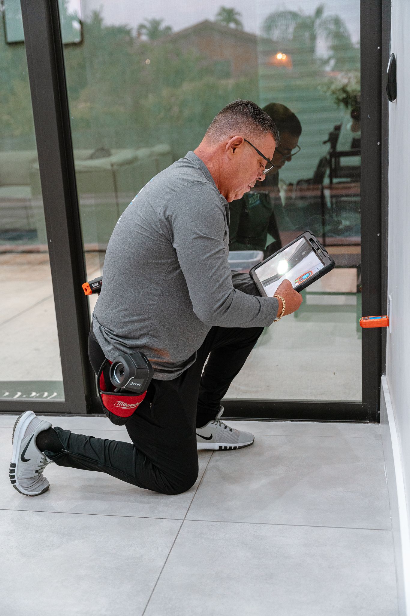 Person kneeling, inspecting a glass door, holding clipboard. Outdoors, light grey clothing, gray and white shoes.