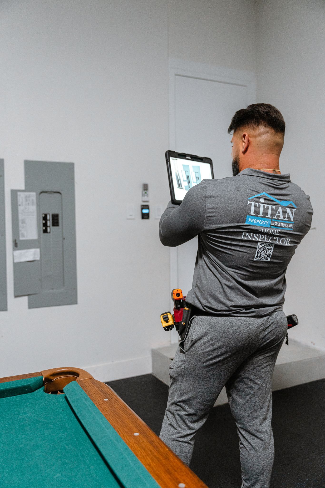 Person in gray sweatsuit holding tablet near a fuse box and pool table.