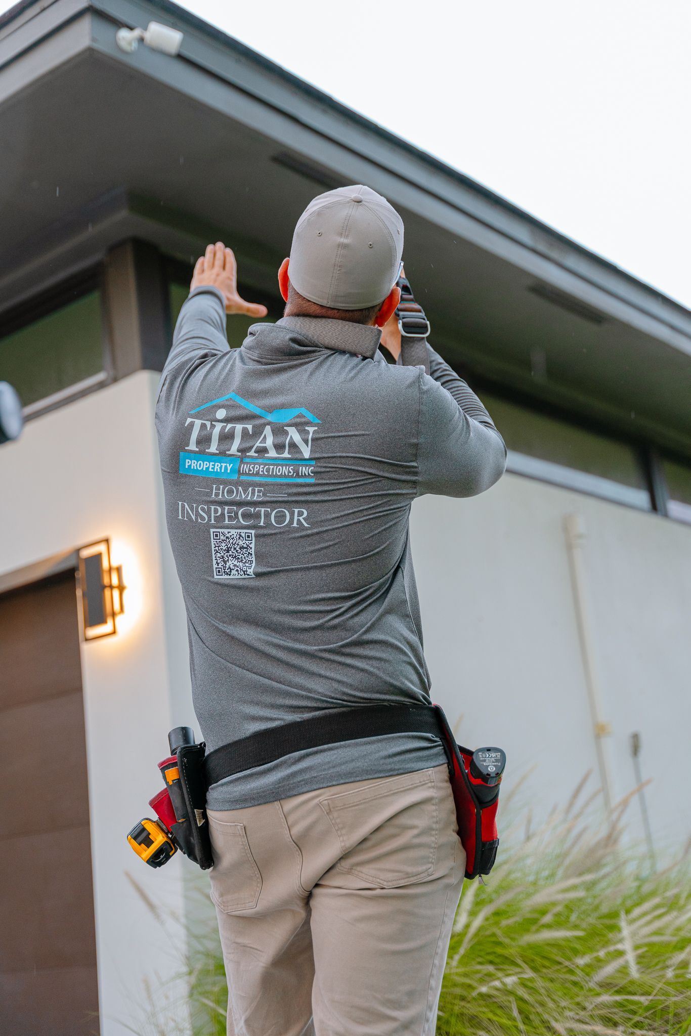 Man in gray shirt installs security camera on a house, tool belt visible.