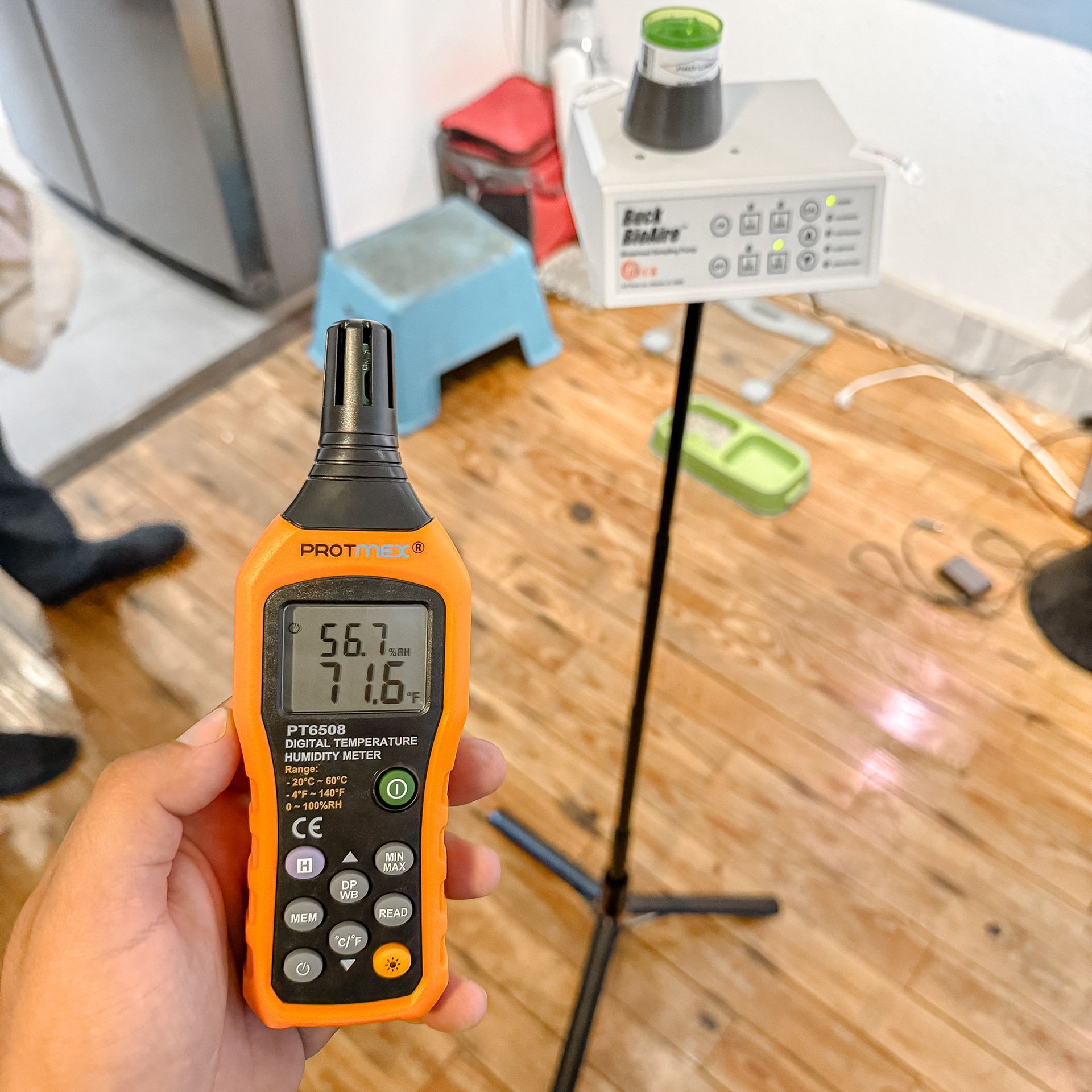 Hand holding an orange air quality meter, reading 56.7 and 71.6, next to an air quality monitor on a tripod.