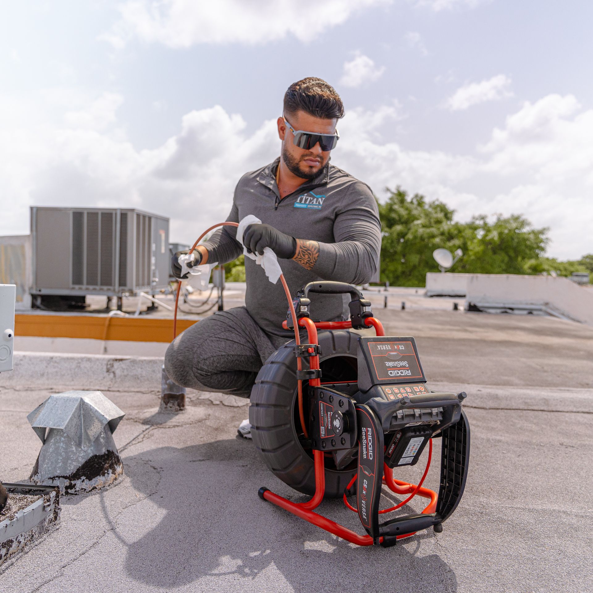Man on a roof operates drain inspection equipment; gray outfit, sunglasses, sunny day.