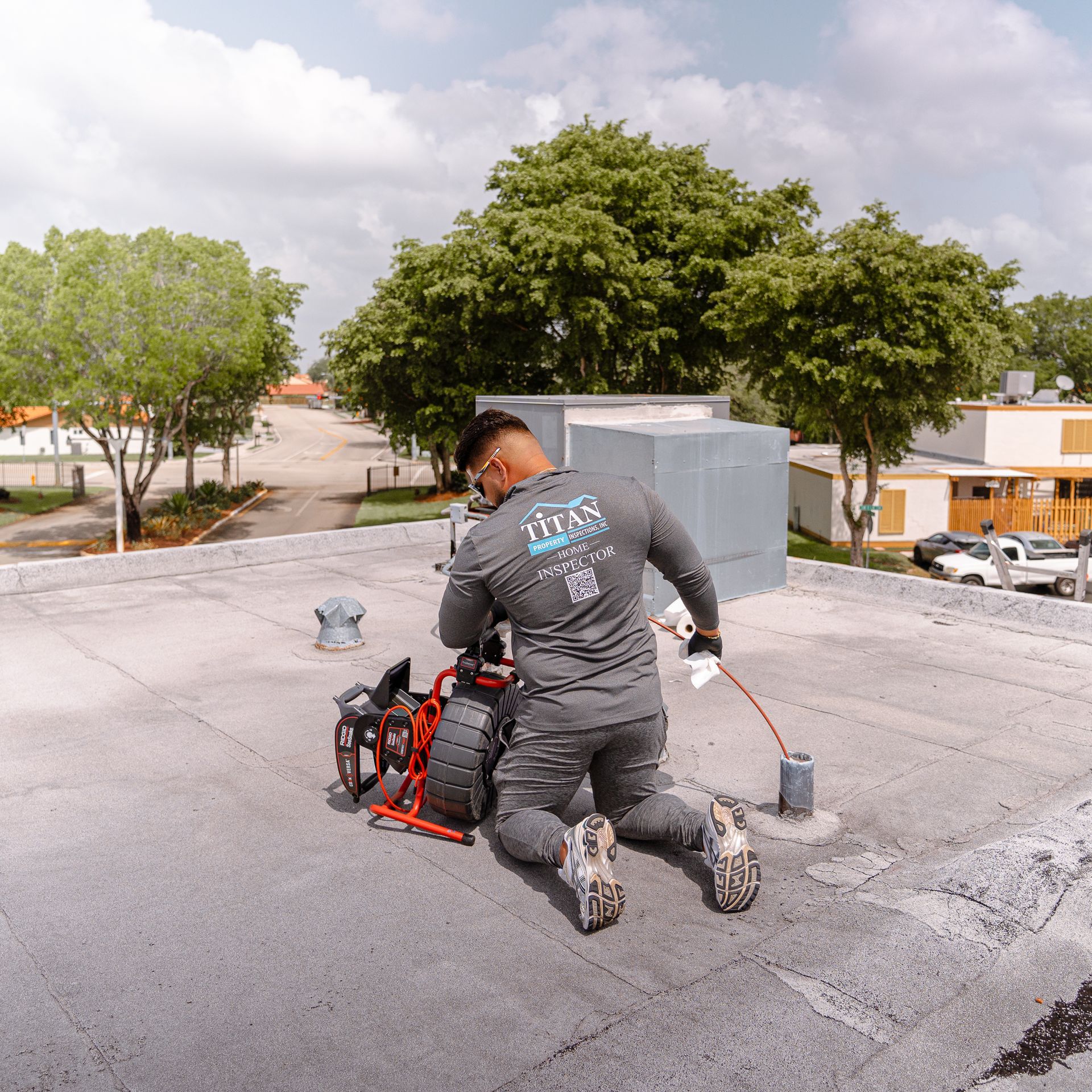A person on a flat roof uses equipment to inspect a drain. The sky is bright, with trees in the background.