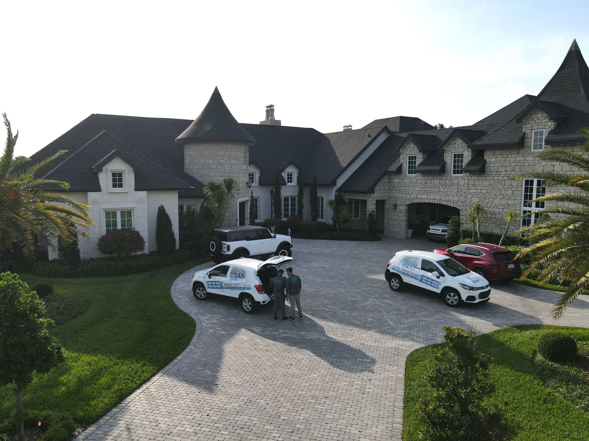 Cars parked at a large stone house; people by one. Lush green lawn, cobblestone driveway. Blue and white service vehicles.