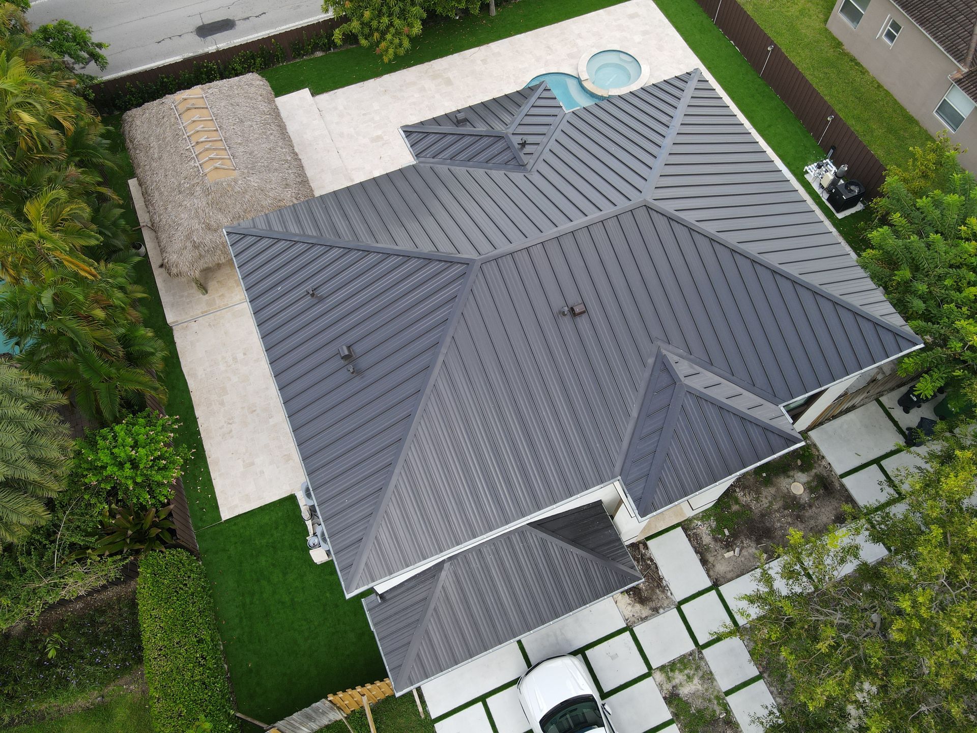 Aerial view of a gray-roofed house with a white driveway, pool, and a tiki hut. Lush green lawn surrounds the property.