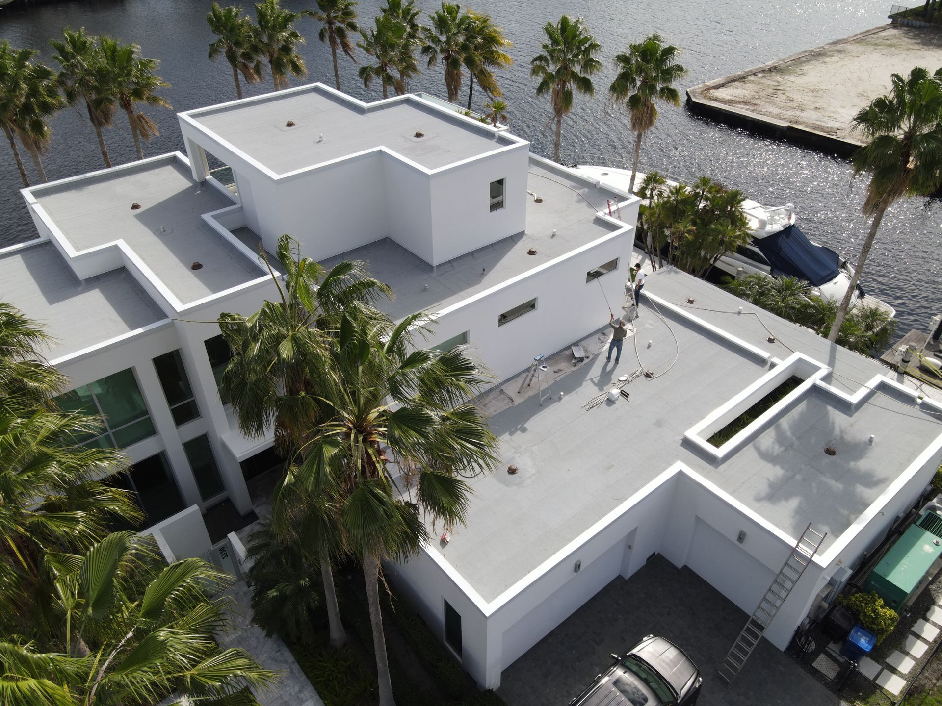 Aerial view of a modern white house with a flat roof, near water and palm trees.