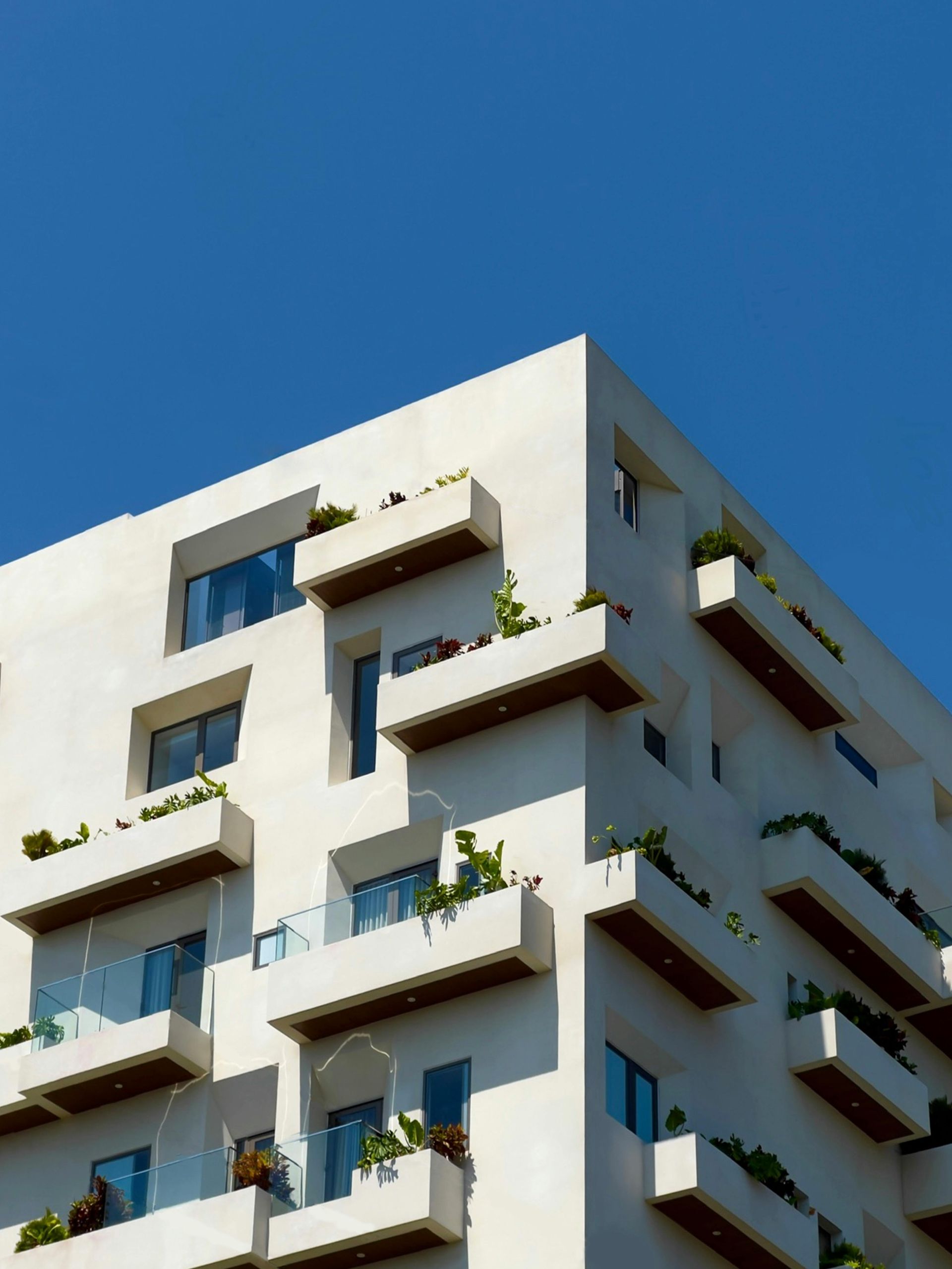 White modern building with angular balconies, lush greenery, and blue sky.