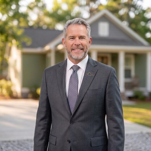 Bernard Oostra in a gray suit standing in front of a house, smiling on a sunny day
