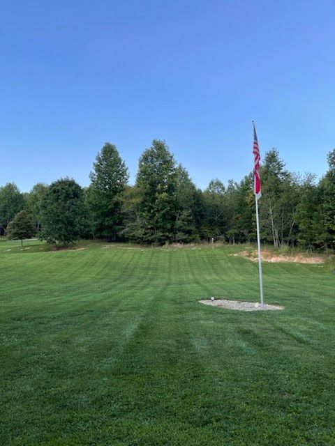 Green lawn with flag on a pole, trees in background, blue sky.
