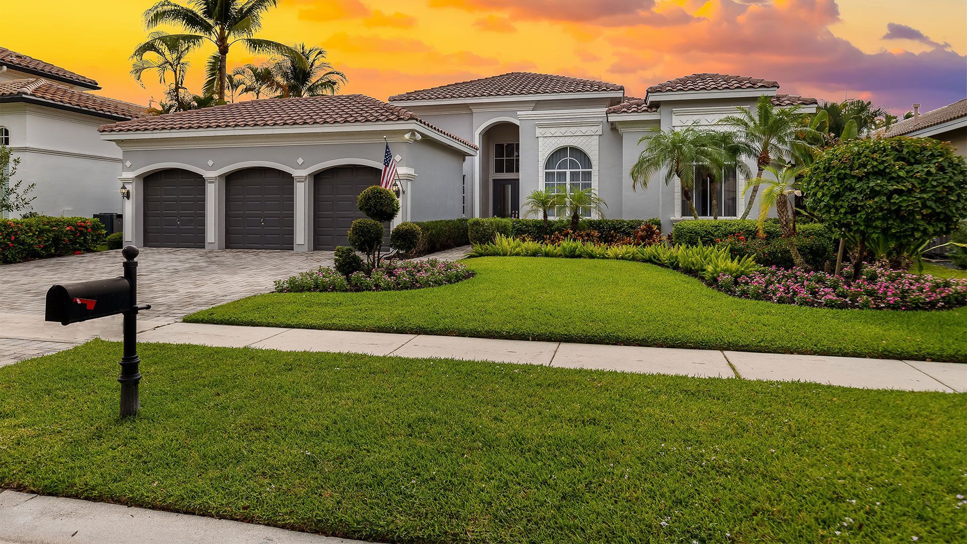 Suburban home with gray facade, three-car garage, lush green lawn, and vibrant sunset sky.