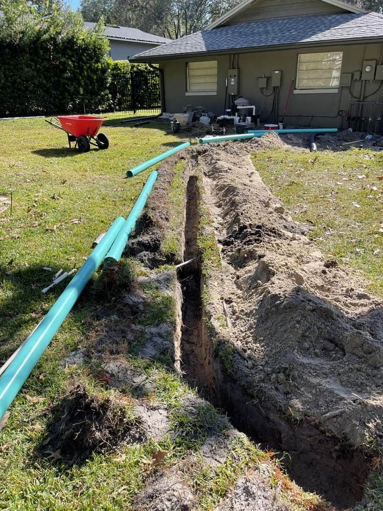 A trench dug in a backyard with blue pipes laid out, likely for plumbing work.