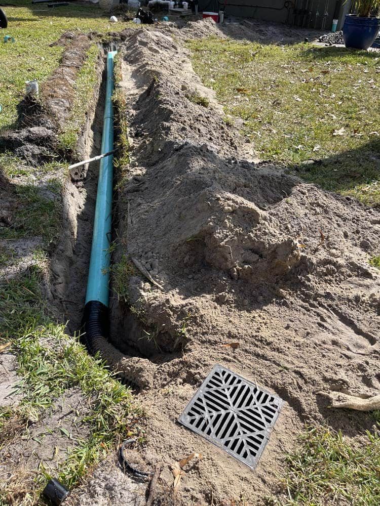 A trench in a yard with blue drainage pipe, a drain grate, and piles of dirt.