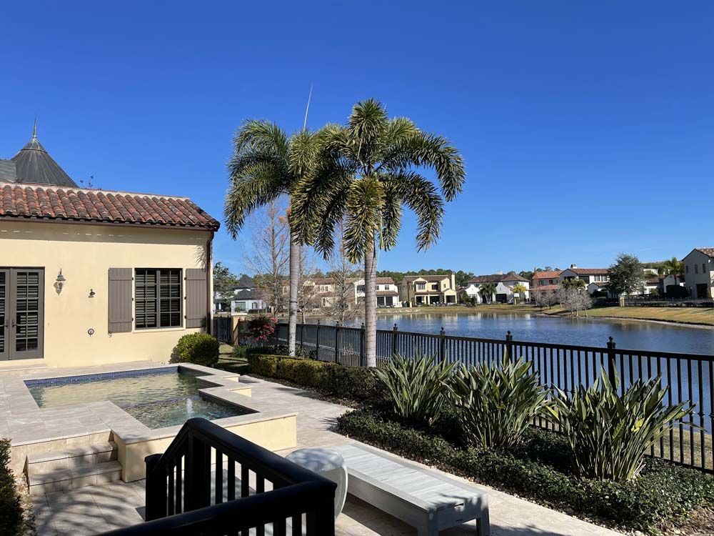 Patio by a lake with palm trees. Tan house, black fence, clear blue sky.