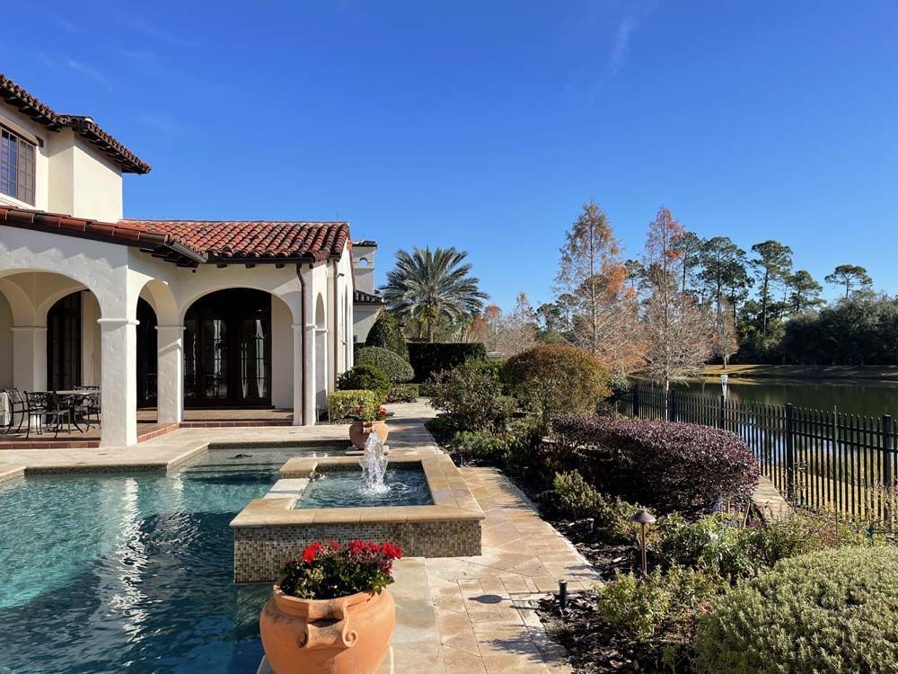 Poolside view of a luxury home with fountain, blue sky, and lake in background.