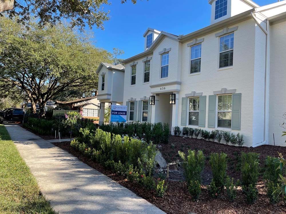 Two-story white building with green shutters, bushes, and a sidewalk on a sunny day.
