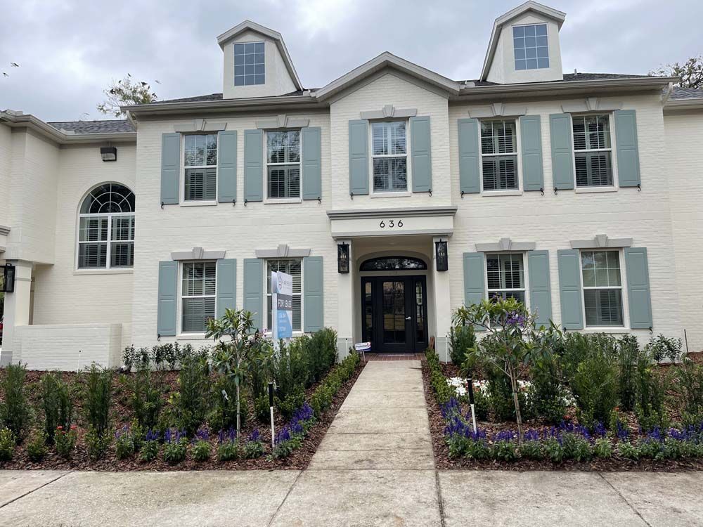 White brick home with light blue shutters and black front door. Small bushes and plants line the walkway.