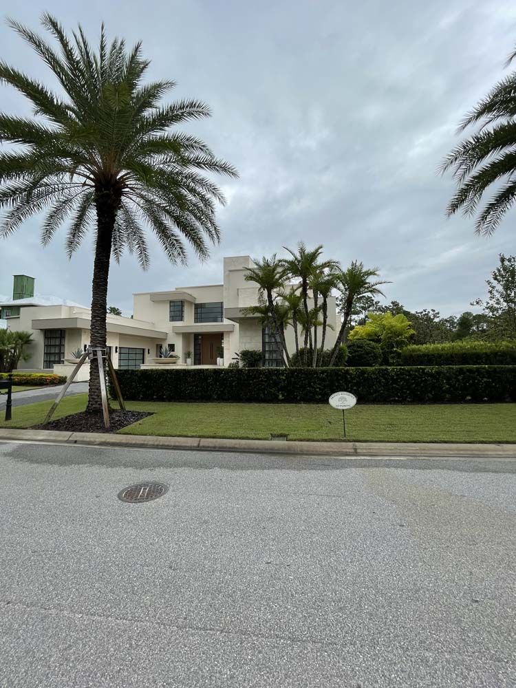 Modern light-colored house with palm trees, green hedges, and overcast sky.