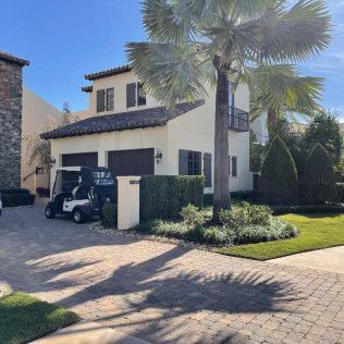 Beige two-story house with dark shutters, garage, and golf cart parked in the driveway. Palm tree in front.