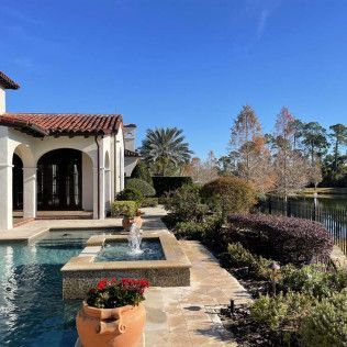 Exterior view of a luxury home with a pool, fountain, and manicured landscaping, next to a lake under a blue sky.