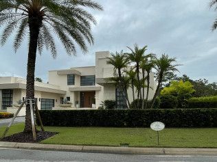 Modern, two-story beige house with palm trees and green lawn, in a residential neighborhood. Cloudy sky.