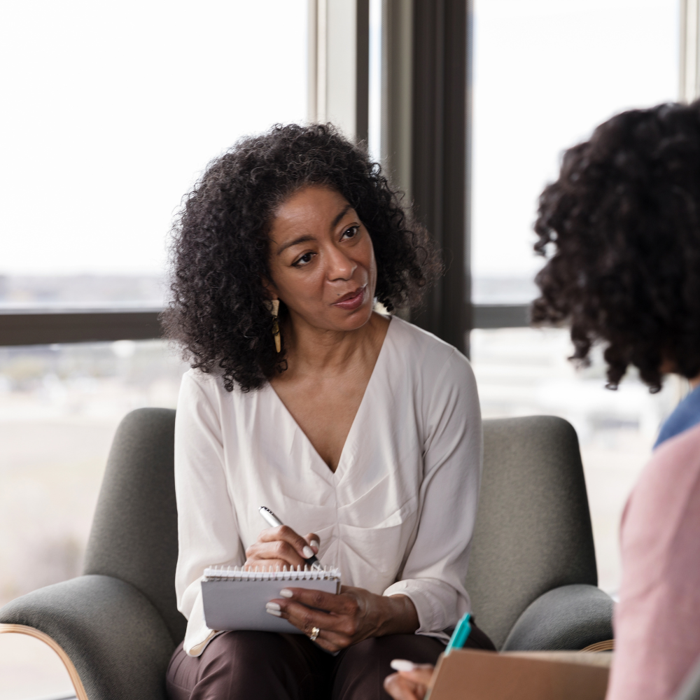 Two people talking in a bright office, one taking notes on a notepad.