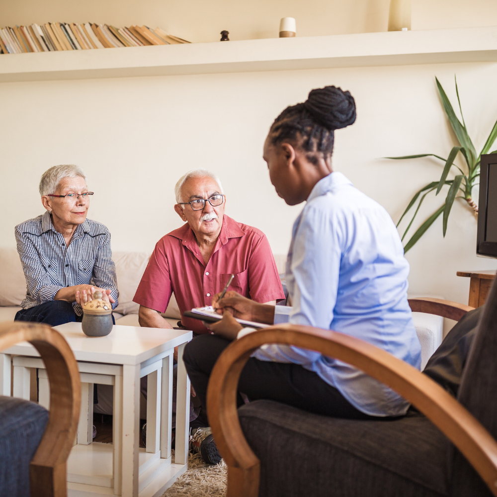 A professional speaks with two individuals in a brightly lit room, taking notes while they sit in armchairs.