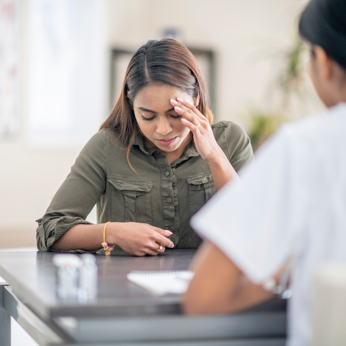 A person sits at a table appearing distressed with a hand on their forehead, meeting with a healthcare professional.