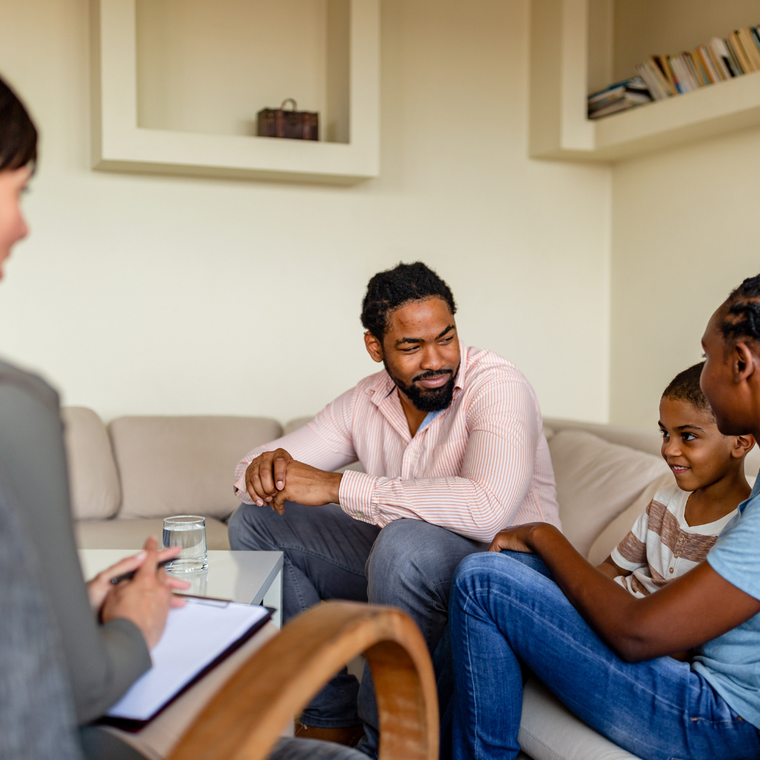 A family sits on a couch in a professional office, participating in a consultation while a provider takes notes.