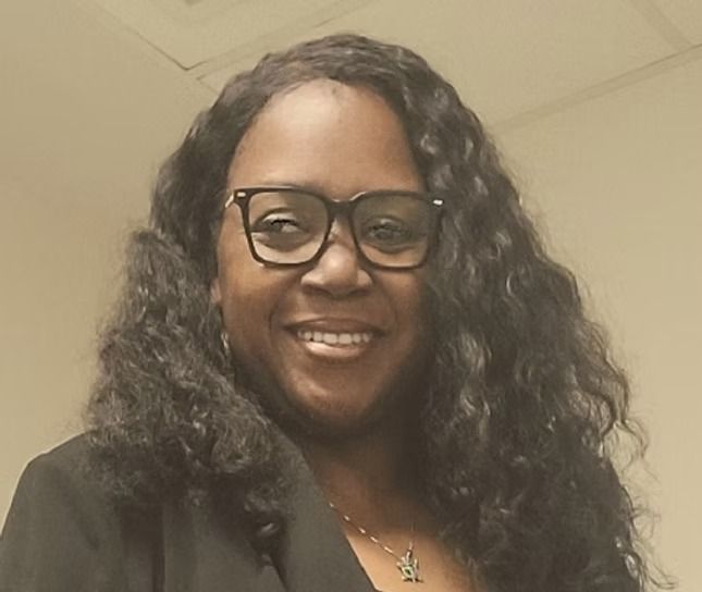 A person with long, wavy dark hair and glasses smiles while wearing a dark blazer and a small pendant necklace.