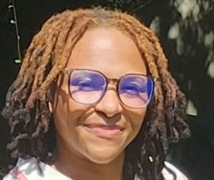 A smiling person with shoulder-length, two-toned dreadlocks wearing glasses against a blurred outdoor background.